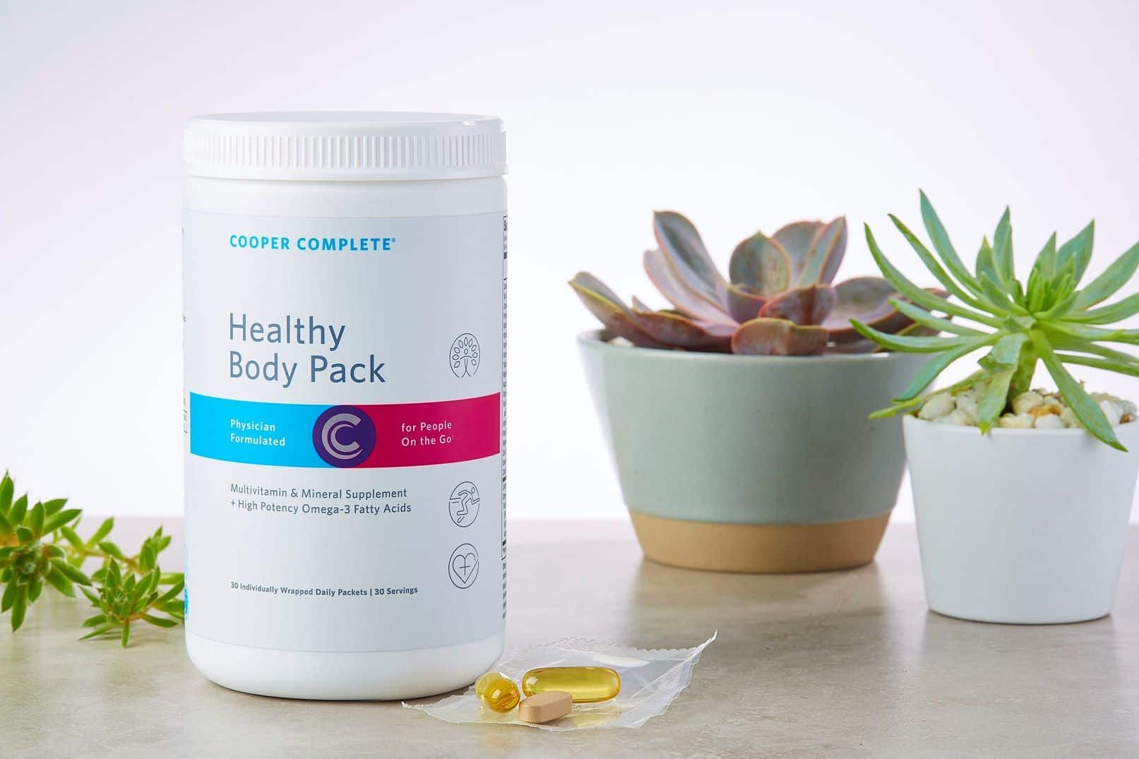 Cooper Complete Healthy Body Daily Vitamin Pack dietary supplement canister and a single daily packet on a counter with several succulent plants