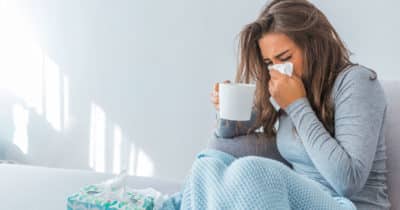 Image of a woman with a cold or flu sitting on a couch bundled up with a blanket drinking a warm drink with a kleenex at her nose