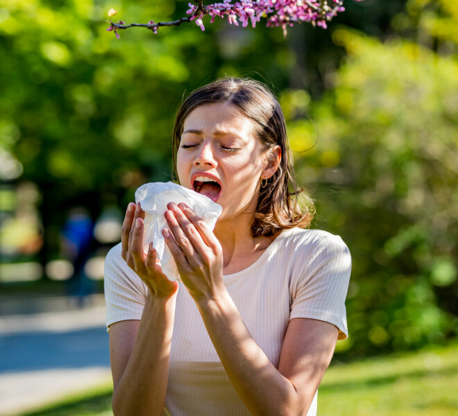 Photo of a woman in a park setting sneezing from seasonal allergies