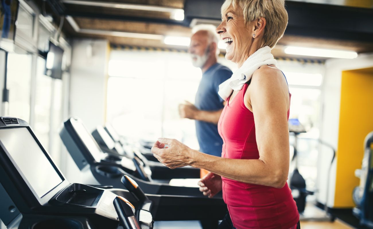Photo inside a gym in the treadmill area with a woman laughing and smiling at someone outside the frame while an older man walks in the background.