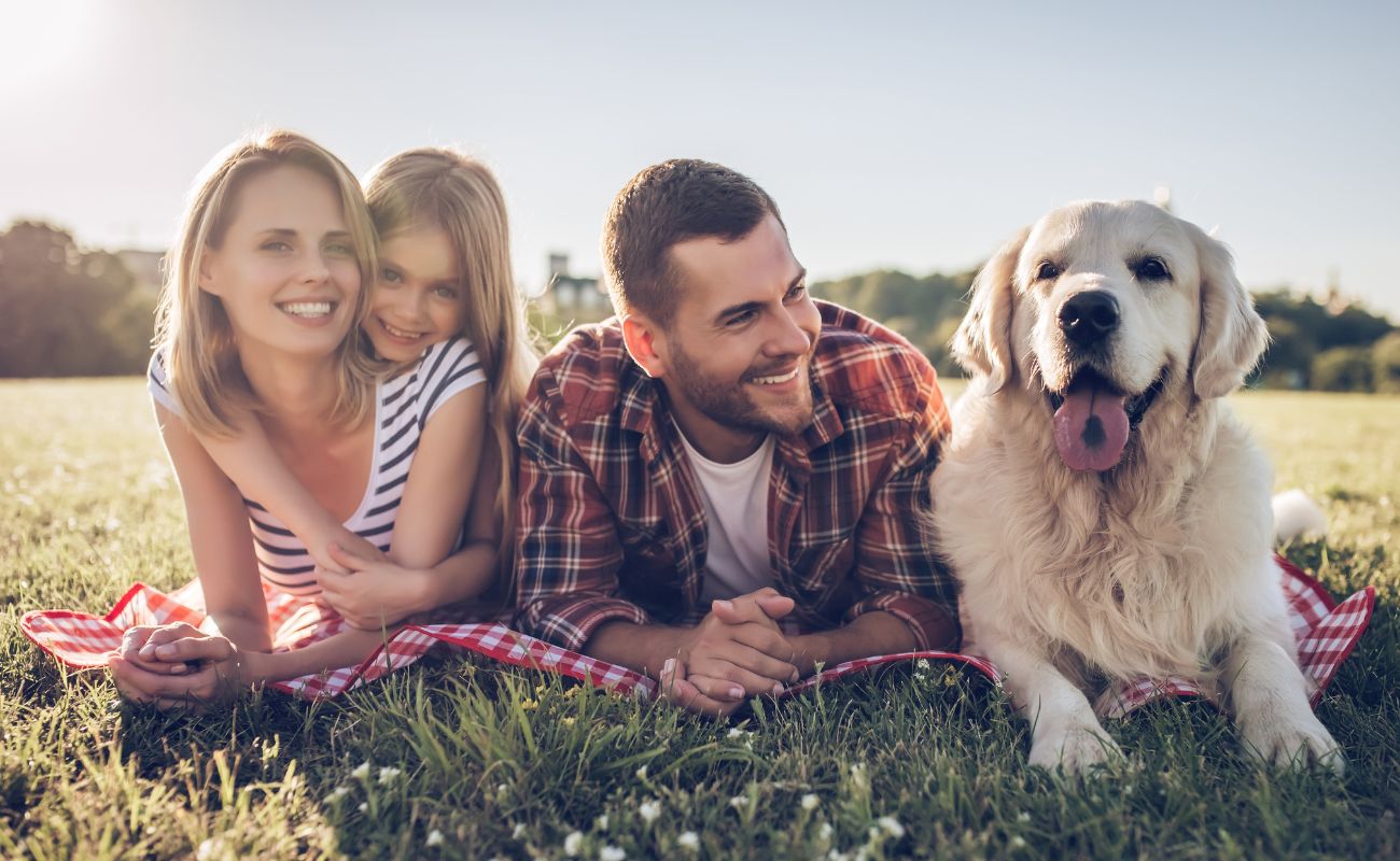 Photo of a young family with their little girl and family dog enjoying the sunshine on a blanket outdoors.