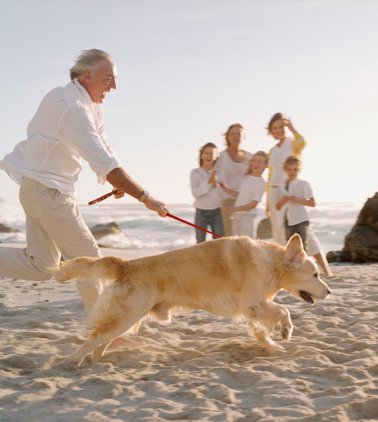 Photo of a multigenerational family enjoying time on the beach as the granddad runs past with the family pup.
