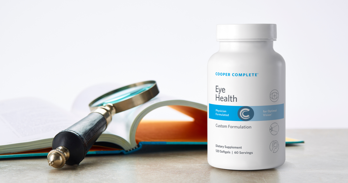 Bottle of Cooper Complete Eye Health dietary supplement on a counter beside an open book and magnifying glass.