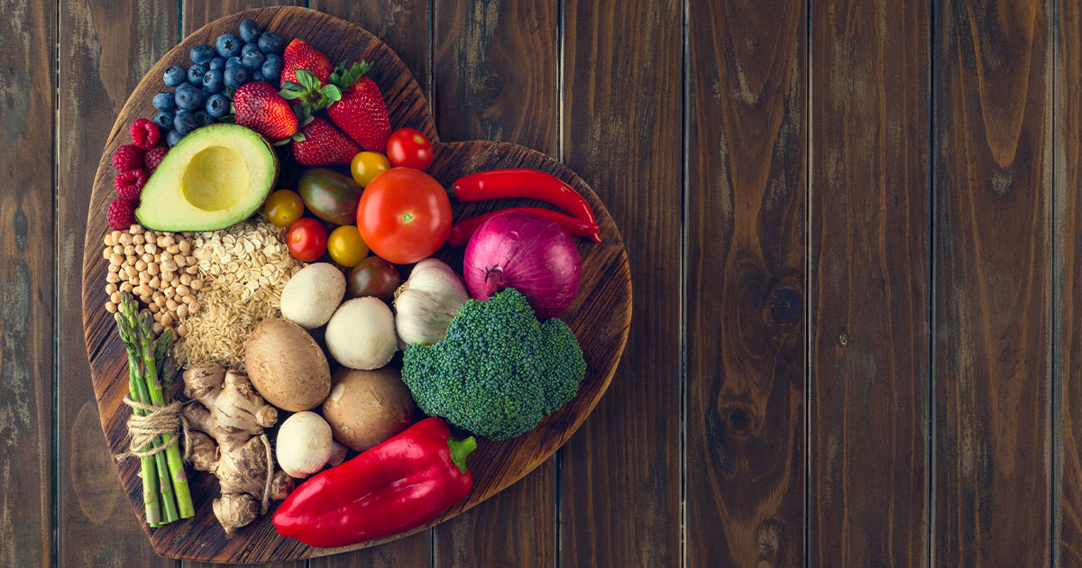 Photo of a wooden heart-shaped bowl of heart healthy vegetables, fruits and whole grains on a brown wooden counter.